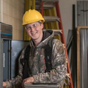 An electrical technology student wears a yellow hard hat and smiles